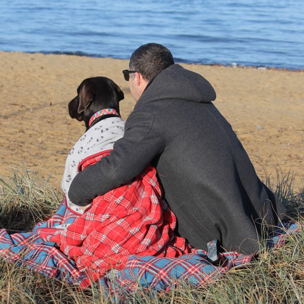 A man and his dog are sitting on the beach looking out to sea. The dog is wrapped in a red check fleece blanket.