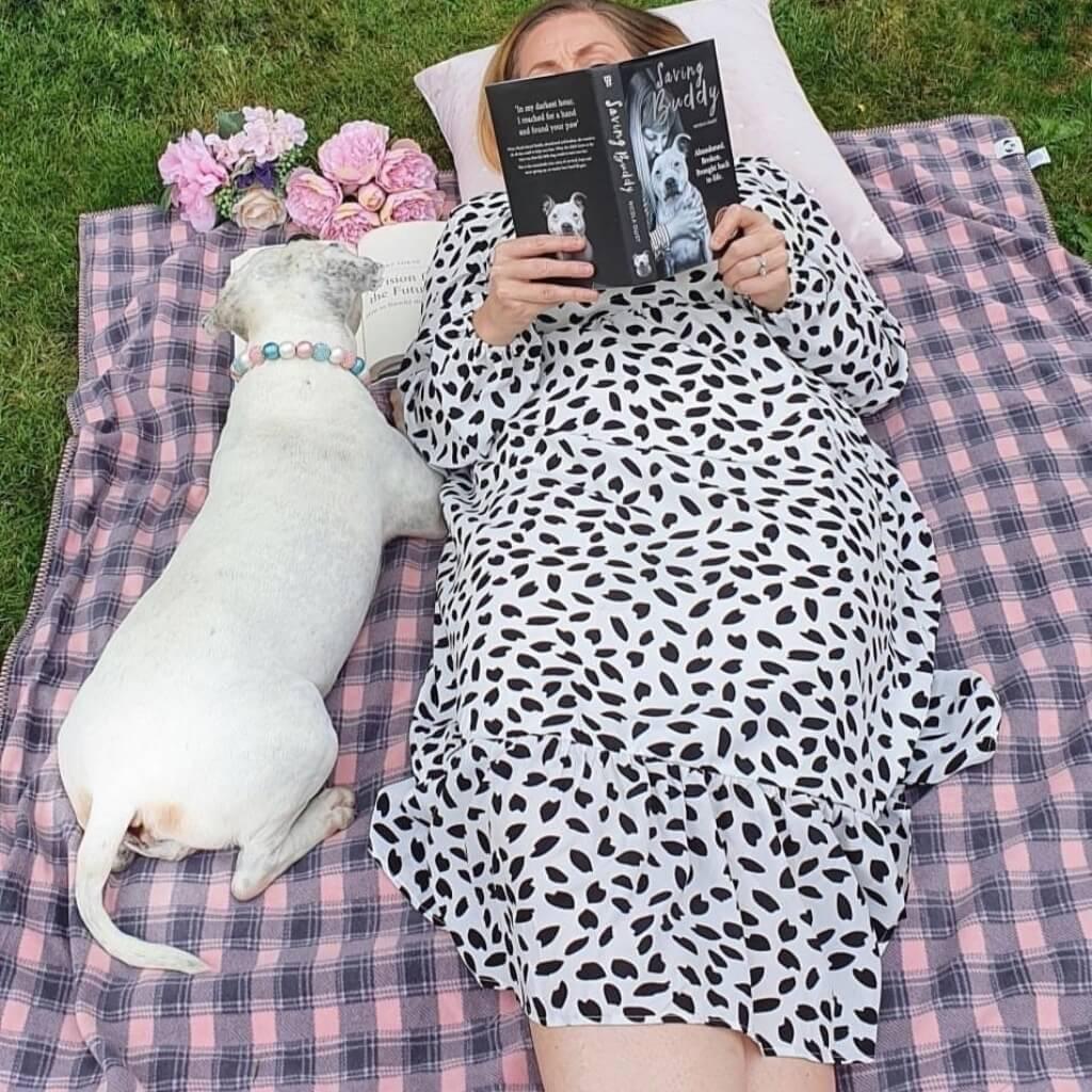 A lady reading a book is lying with her dog on a pink check fleece blanket.