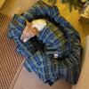 A small brown and white dog is lying in his dog bed which has been covered in a green and blue check fleece blanket.